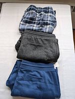 Three pairs of men's pants stacked, displaying the plaid, gray, and blue pants.
