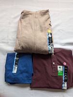 Three folded brand new men's tops in beige, blue, and maroon colors each with attached tags showing size 3XL.