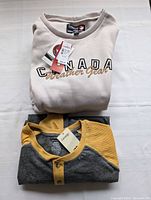 Two folded men's tops, one cream Canada Weather Gear sweater and one gray and mustard yellow long sleeve shirt with buttons.