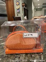 Photo of one of the orange semi-circular makeup bags packaged in clear plastic with a handle on top and some makeup brushes visible.