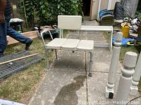 Front view of a used white bath bench with padded backrest, three padded seating areas including a leg rest, metal frame, and adjustable legs.