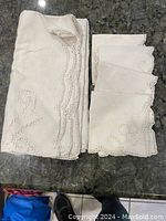 Folded cream tablecloth and six matching napkins with embroidered scalloped edging on a dark surface.