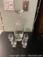 Crystal decanter bottle with stopper and four shot glasses displayed on black surface, showing overall condition and shape.