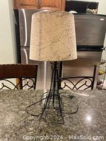 Black metal lamp shaped like Eiffel Tower with beige fabric shade printed with black handwritten script text. Lamp placed on a kitchen counter with chairs in background.