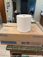 Large single roll of white industrial paper towels displayed on a cardboard box, unopened but removed from packaging to show size.