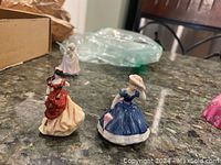 Two mini Royal Doulton porcelain figurines on a countertop. One dressed in a blue gown with a hat covering her face, the other in a red and cream dress missing an arm.