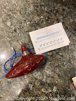 Photo of the red 12-hole ceramic ocarina on a granite countertop alongside the fingering chart and blue neck strap.