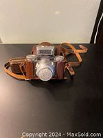 1950's Paxette camera in brown leather carrying case with strap placed on black surface, showing front view of camera and case.