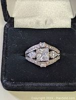 Front view of 925 sterling silver ring with a square princess-cut cubic zirconia stone and multiple smaller round stones on split shank band, set inside a black velvet ring box