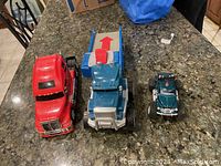 Three toy trucks on a dark countertop: red semi-truck, blue semi-truck, and small green jeep-like truck