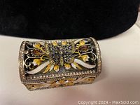 Front angled view of gold-tone trinket box with butterfly and Swarovski crystals on lid, detailed floral filigree sides.