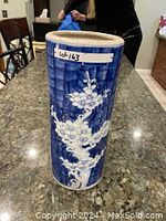 Photo of a tall cylindrical blue and white vase with painted white blossoms on branches against a blue geometric background, placed on a granite countertop indoors.