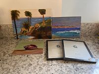 Three paintings and two empty frames on countertop: coastal palm trees, coastal wave scene signed B. Parks, small sea shell painting, two frames.
