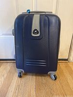 Front view of blue hard shell carry-on suitcase showing logo emblem and textured surface with wheels.