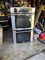 Electrolux double oven stainless steel unit with two glass door ovens, visible handles, placed on a garage floor surrounded by various storage items.