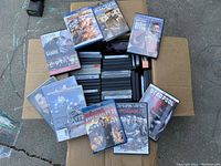 Box containing around 80 DVDs, some new in shrink wrap, others opened. Titles visible include action and western films, Deadpool and The Expendables 2.