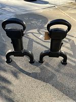 Photo of two black charcoal finish fireplace andirons standing side by side on concrete ground, showing front and side profiles under daylight.