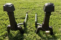 Pair of antique fireplace andirons standing on grass showing front structure and side profile