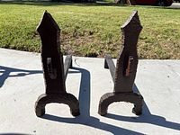 Pair of black metal andirons shown standing upright on concrete with grass in background.