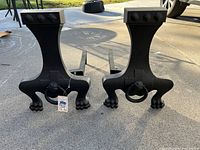 Photo showing a pair of black cast iron fireplace andirons with paw-shaped feet, ring handles and rectangular top bars, placed on a concrete patio surface.