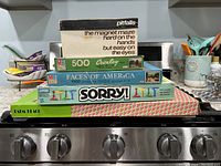 Stacked board games and puzzles including Pitfalls, Sorry!, Palm Beach, Crosley 500-piece puzzle, Faces of America 1000-piece puzzle, photographed on a stove.