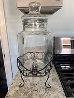 Hexagonal glass beverage dispenser with metal spigot on black metal rack stand, shown on kitchen counter.