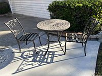 Full shot of outdoor metal patio set on concrete, showing table and two chairs with decorative metal designs and dark finish.