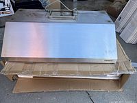 Front view of the stainless steel over range hood on top of a cardboard box in a garage or warehouse setting.