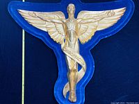 Full view of gold-tone, winged male figure cast metal sign with 'CHIROPRATIC' sash on blue velvet background with measuring tape showing width.