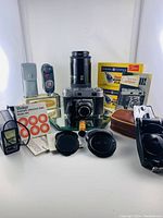 Main assortment of camera and accessories including Kodak Retina III C camera, flash units, flashbulb boxes, remote, and light meter spread on white surface.