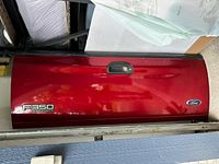Red Ford F350 tailgate with emblems and handle, laying against wall, showing front of the tailgate.