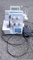 Front-right side view of white Baby Lock BL4-736DF serger with power cord and foot pedal on asphalt surface