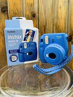 Front view of the blue Fujifilm Instax Mini 9 instant camera next to its boxed packaging, showing camera body, flash and wrist strap.