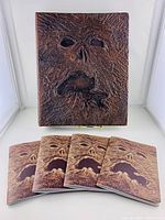 Main Necronomicon book replica with textured, grotesque face cover displayed on stand with four booklets in front showing the same cover art.