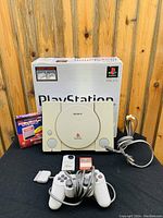 Front view of Sony PlayStation 1 console with controller, memory card, game cartridge, and original box in background.