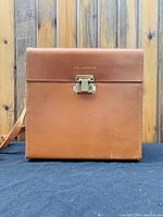 Closed brown leather Polaroid carrying case photographed from front on a black surface with wood background.