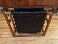 Front and side angled view of teak frame and black leather sling magazine holder on wooden floor with mid century furniture in background.