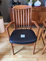 Front view of teak arm chair showing vertical slat backrest, armrests, and black upholstered seat.