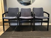 Front view of 3 black painted teak arm chairs with gray cushions arranged side by side against a wall with a mirror above.