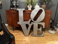 Full view of large metal LOVE sculpture, showing all four letters arranged with O tilted, placed on wooden floor in front of wooden sideboard.