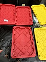 Five large 27 gallon plastic storage totes with red and yellow lids stacked open showing example contents inside one tote.