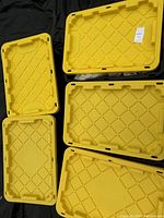 Five yellow plastic 27-gallon storage tote lids with diamond textured pattern, arranged on black surface, showing different angles and tops of lids.
