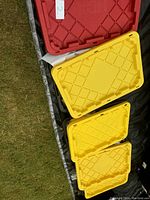 Four storage tote lids stacked on grass showing red and yellow lids with diamond grid pattern and label '2' on the red lid.