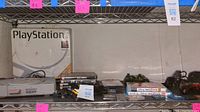 A shelf with an original PlayStation box, a gray PlayStation console, several PS2 game cases, multiple controllers, and cables.