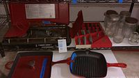 Photo showing Century Primus gas stove with lid open, red Mario Batali cast iron pan and Nutrichef grill pan in front, along with knife set and glass jars.