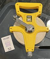 Top view of yellow Power Fist branded tape measure reel with white 100 ft tape and metal winding handle