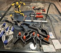 Photo showing 18 various clamps on a glass table, including quick-release grip clamps, spring clamps, and screw clamps.