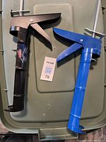 Top view of two manual caulking guns, one black and one blue, positioned side by side on a plastic surface with a lot tag.