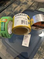 Three rolls of tape on glass table: green Safety Caution Tape, yellow Stanley Painters Tape, and silver Foil Tape