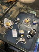 Seven assorted padlocks with keys placed on a glass tabletop. Two brass-colored locks, two Master brand locks (including a blue and silver combination lock), one rusted round lock, and other smaller locks visible with keys and tags.